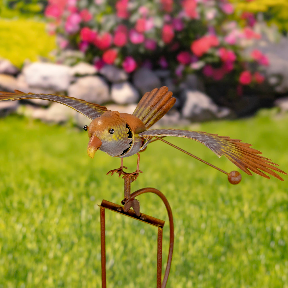 Close view of the bird’s face and spread wings above its curved stake, with flowers and lawn behind it.