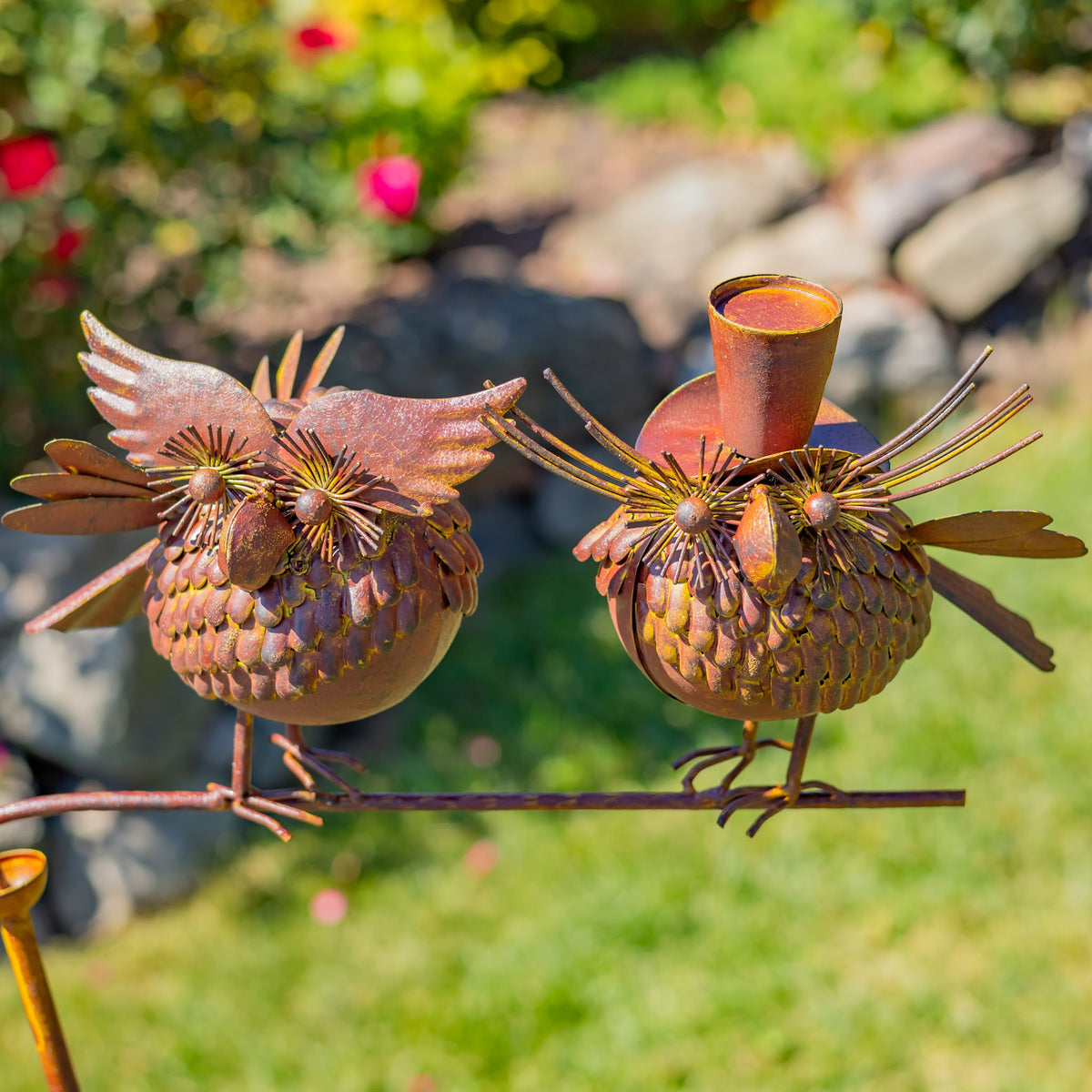 Close view of two decorative metal birds balancing on a rod, with grass and a stone path behind them.
