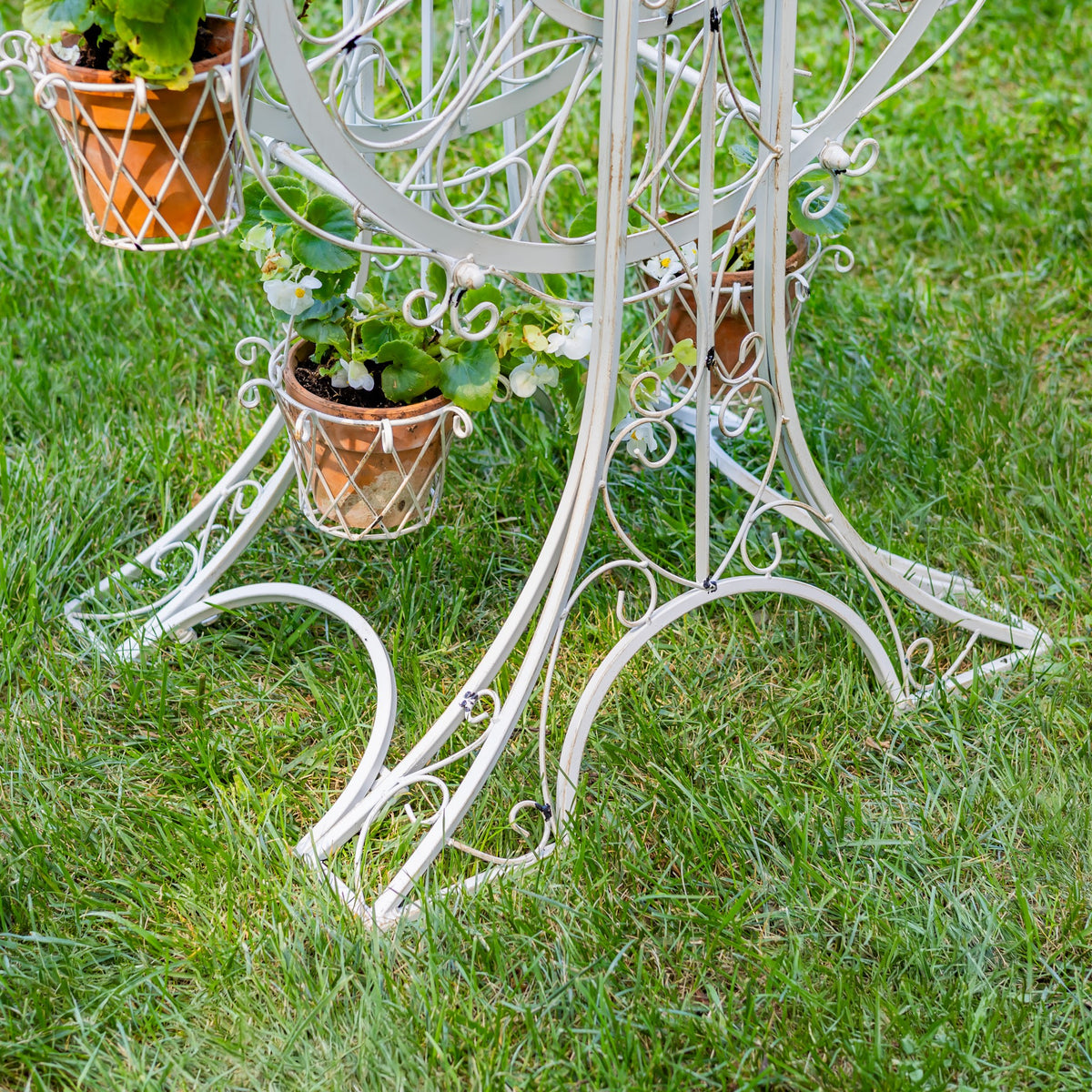 Curved white metal legs and scrollwork support terracotta-colored baskets filled with flowers on grass.