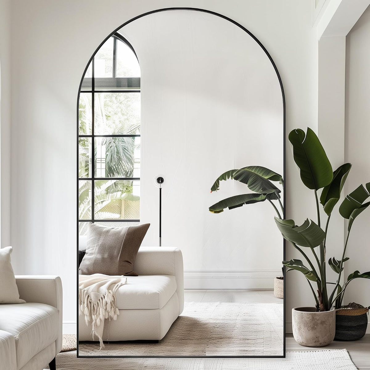 Oversized arched floor mirror with thin black frame standing on the floor, reflecting a sectional sofa, window, and potted