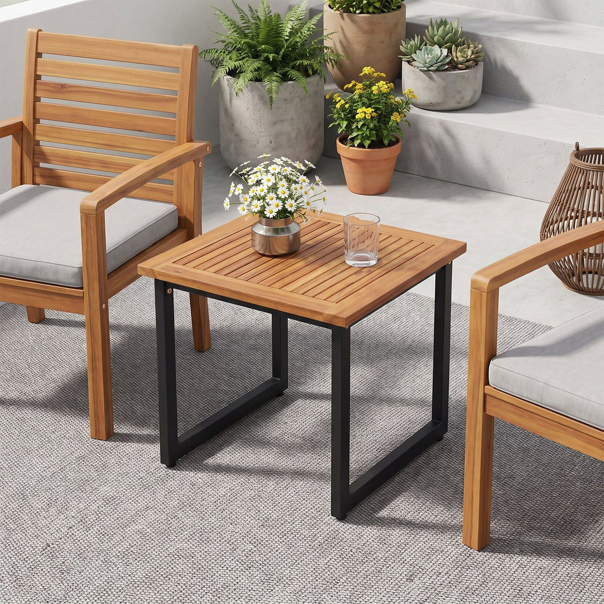 Square slatted wood table with dark metal legs between two cushioned chairs, surrounded by potted plants.
