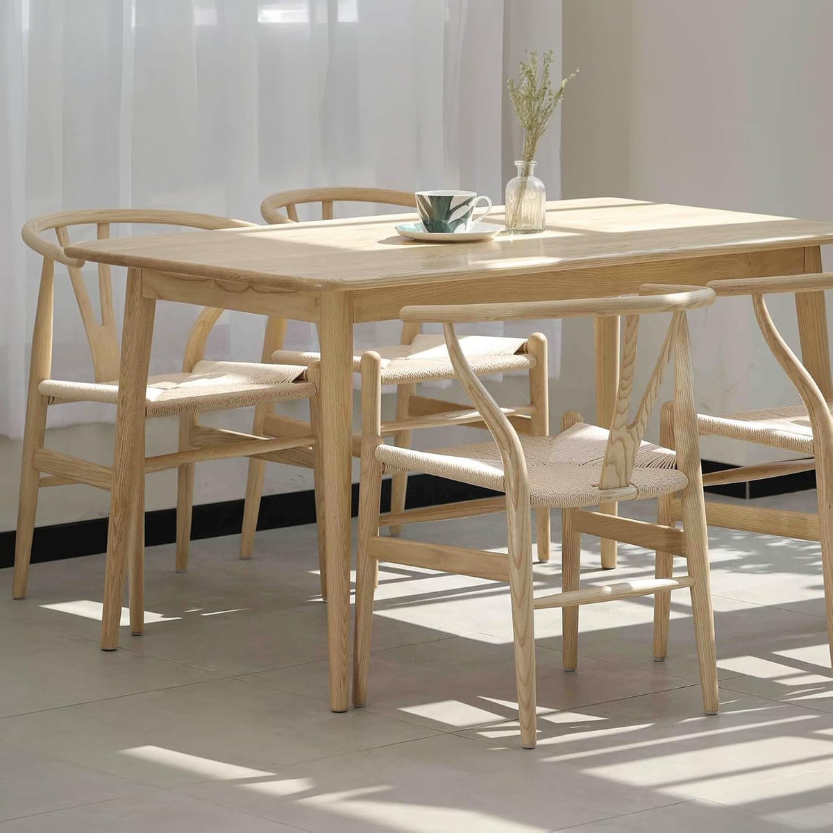 Dining scene with a light wood table and three wishbone-style chairs in sunlight by a sheer curtain.