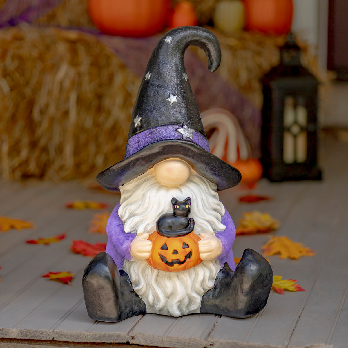 The witch-hat gnome sits on a wooden surface amid blurred Halloween decorations, holding a pumpkin with a black cat.
