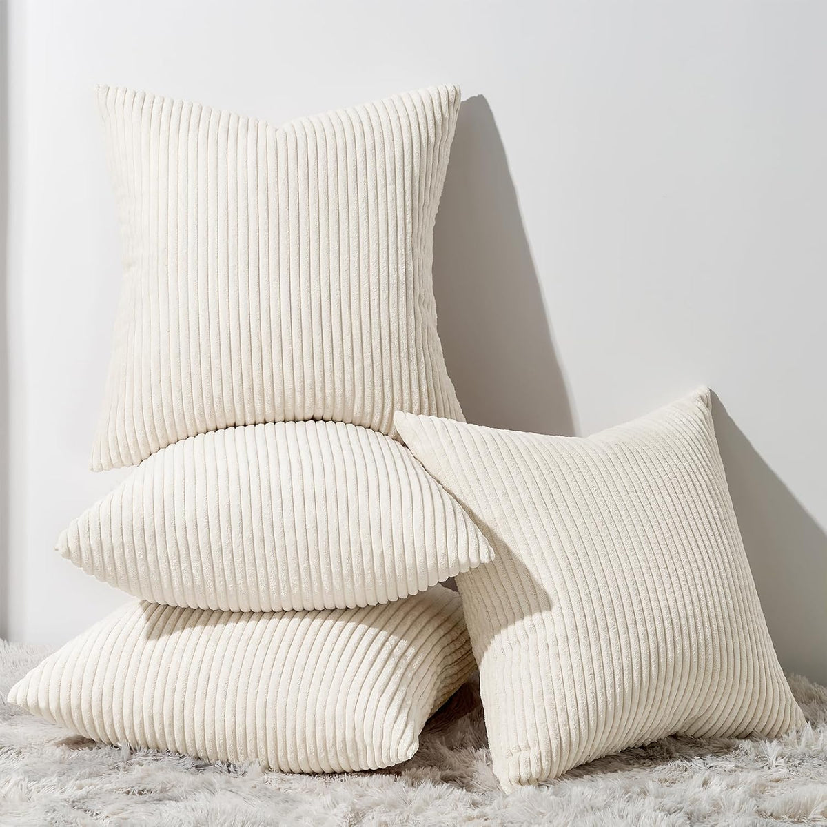Stack of three cream ribbed square pillows on a fluffy rug against a white wall.