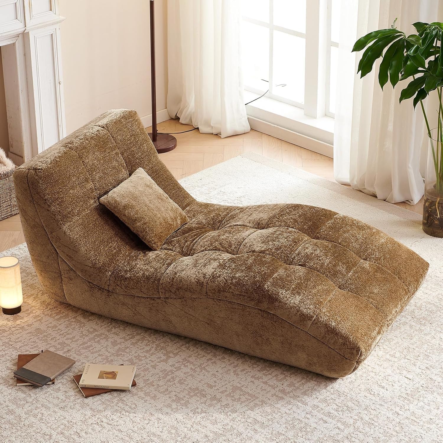 Angled overhead shot of a curved tufted chaise in golden-brown plush fabric with a matching pillow, lamp, books, and a