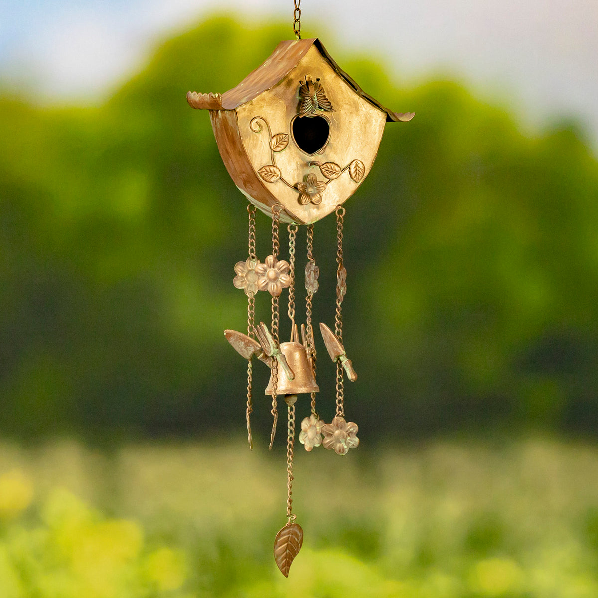 Decorative hanging birdhouse with a heart-shaped opening, flower details, chains, and a small bell against greenery.
