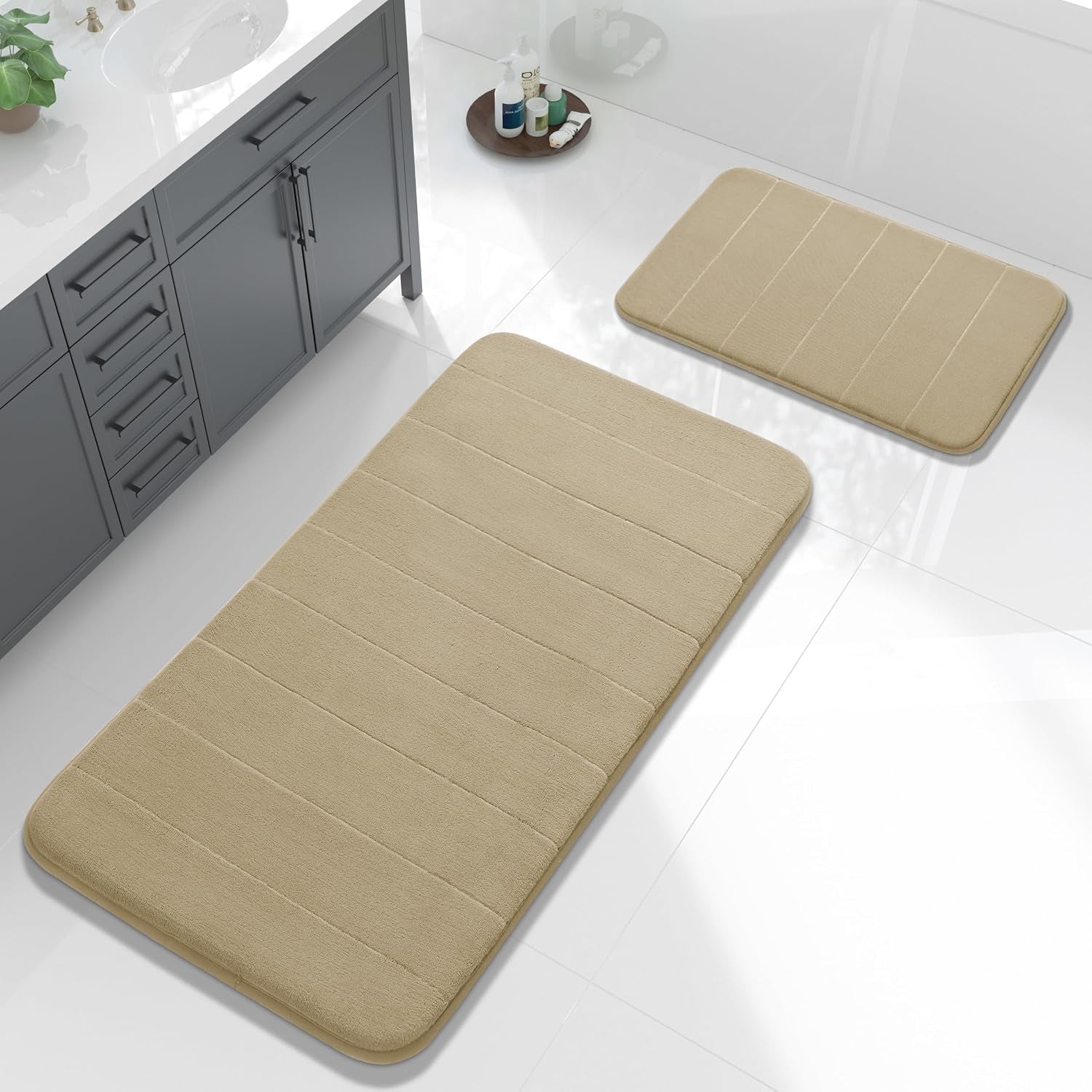 Two tan/beige bath mats arranged in front of a gray vanity on a bright tiled floor.