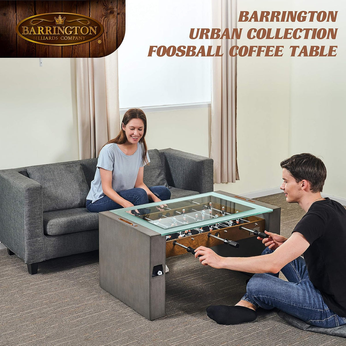 Two people play on a glass-top foosball coffee table in a living room with a gray sofa and beige curtains.