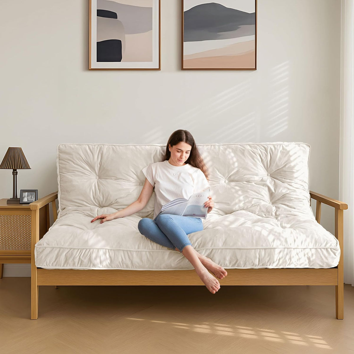 Cream tufted futon mattress on a light wood frame with a woman reading in bright natural light