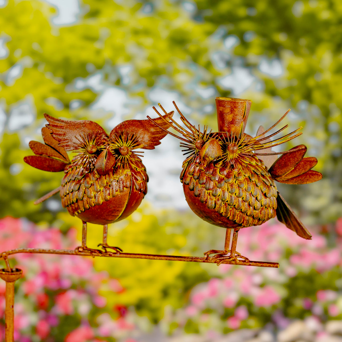 Two rust-colored metal birds balance on a horizontal rod against a leafy garden background.