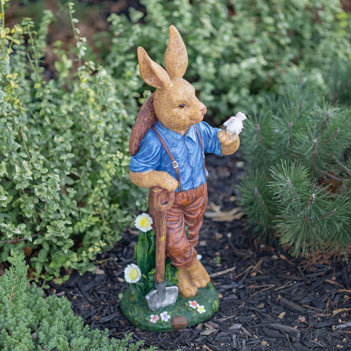 Angled view of the rabbit statue holding a bird and shovel, standing on a flower-painted base in a garden.