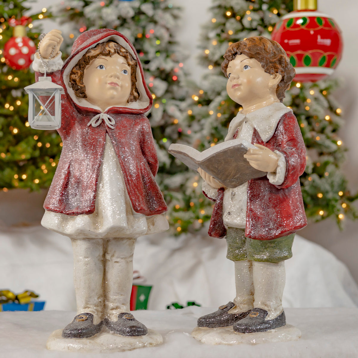 Two childlike Christmas figures in red cloaks stand together, one holding a lantern and the other an open book.