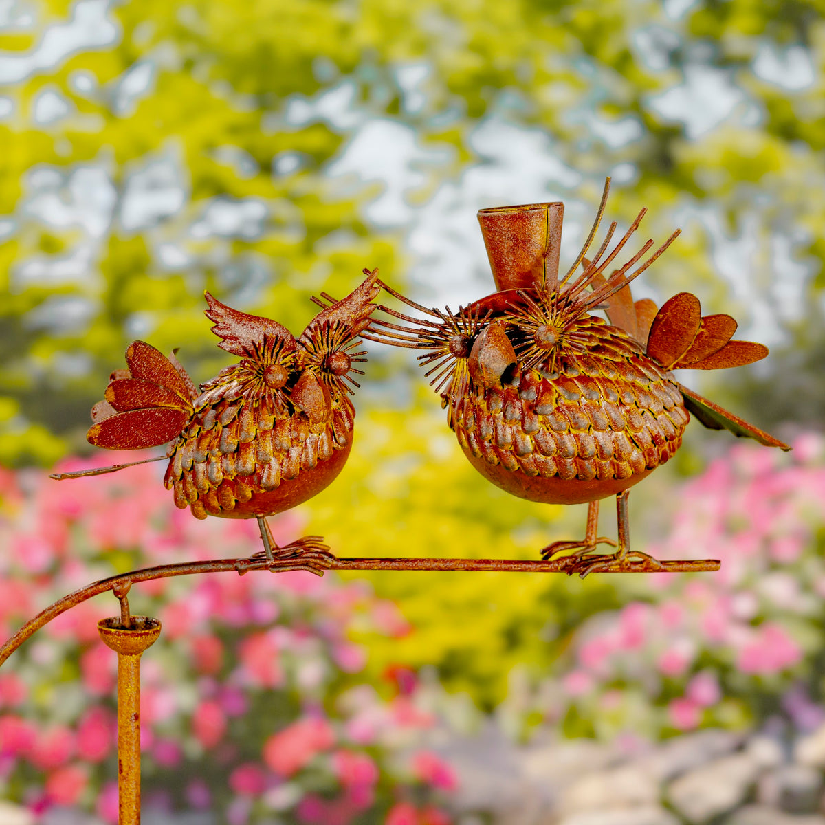 Two rust-colored metal birds balance on a horizontal garden stake against a blurred flower garden.