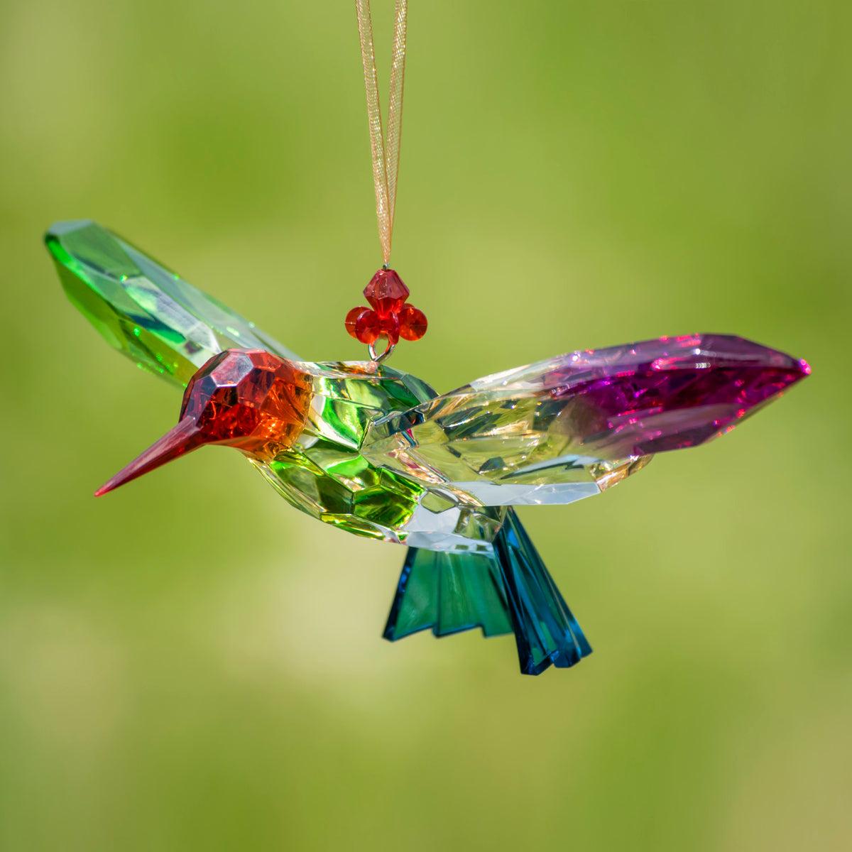 Multicolored faceted hummingbird ornament hanging against a soft green background, with a red-orange head and blue tail.