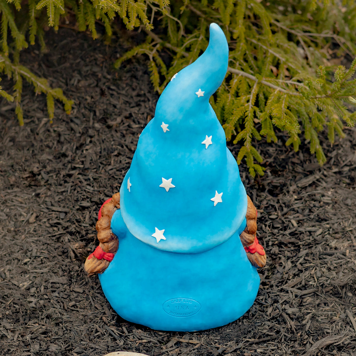 The gnome’s blue star-covered hat and rounded back are shown among garden mulch and greenery.
