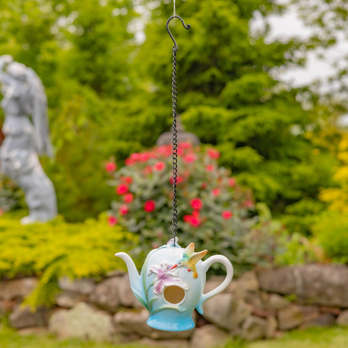 Small teapot-shaped birdhouse suspended from a long chain in a garden, with a stone border and greenery behind it.