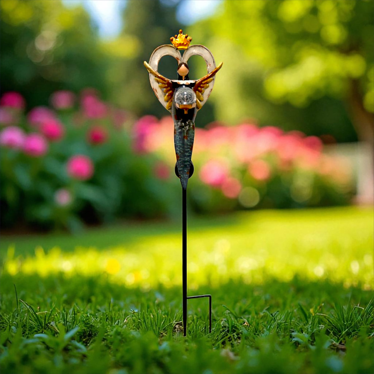 A front-facing angel stake with raised wings and a crown stands in grass against a colorful garden.