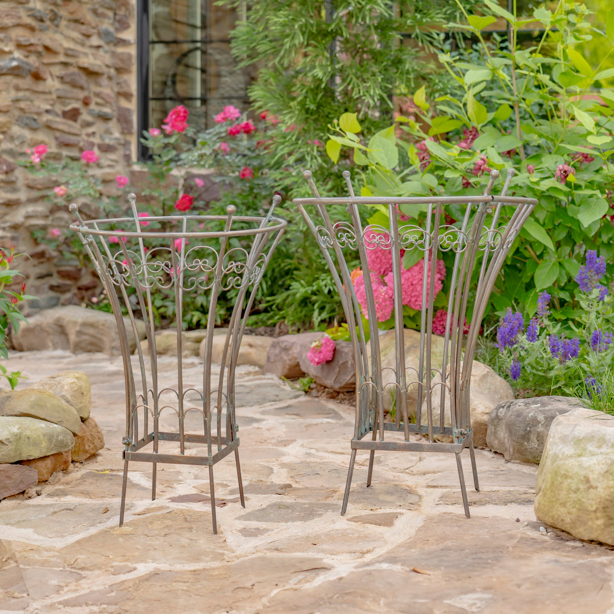 Two round iron trellis stands on a stone patio among flowering plants.