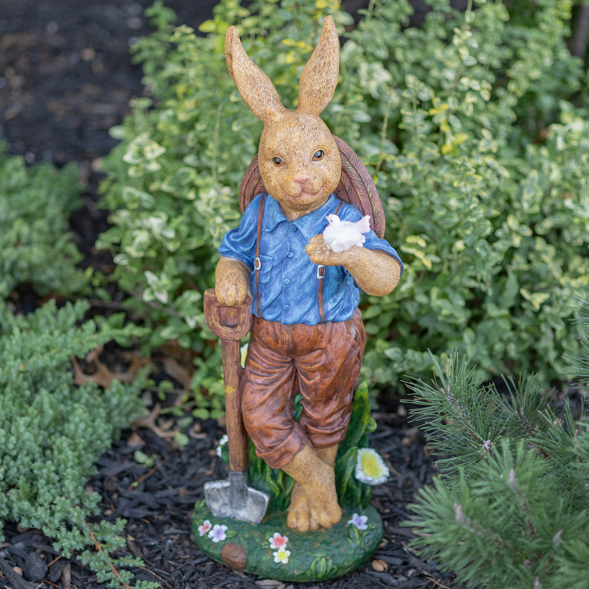 Rabbit statue in a blue shirt and brown trousers holding a white bird and shovel among garden plants.