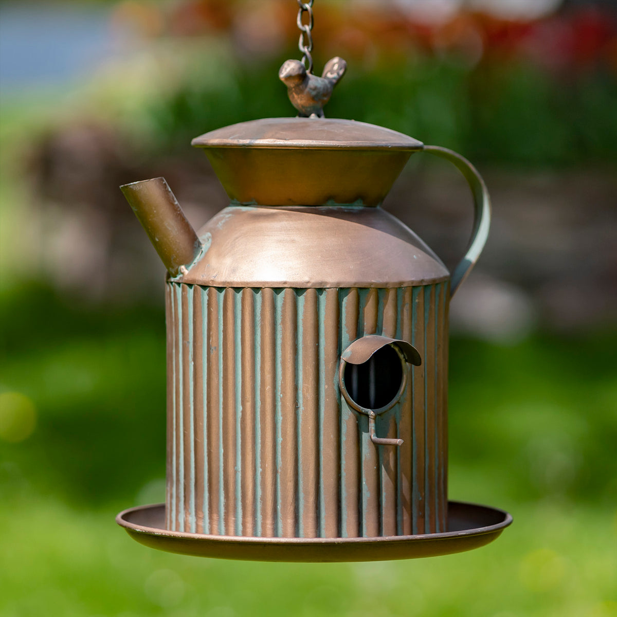 Antique copper-colored teapot-shaped bird feeder hangs against a blurred green garden background.