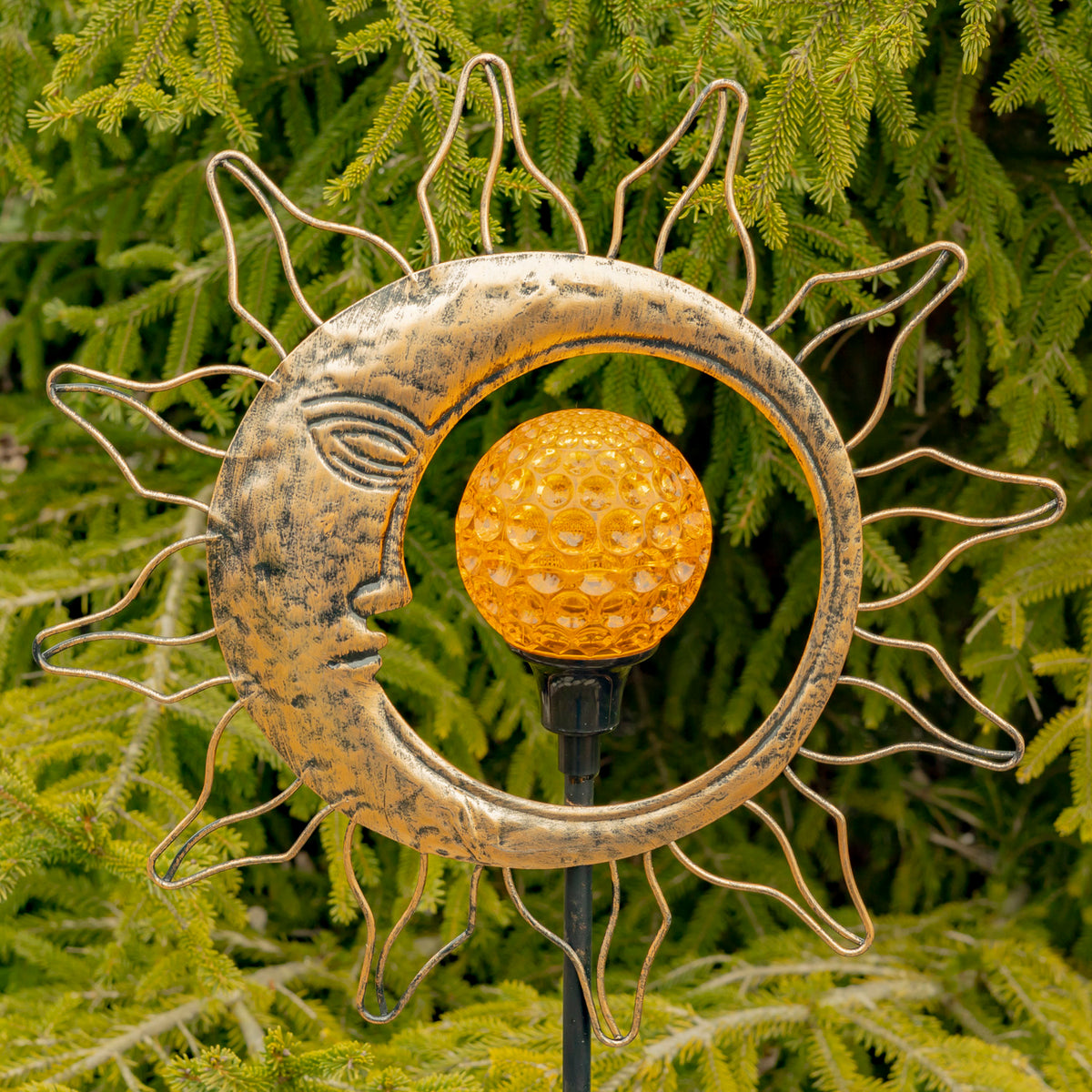 Gold sun-and-moon garden stake with an amber globe among evergreen branches.