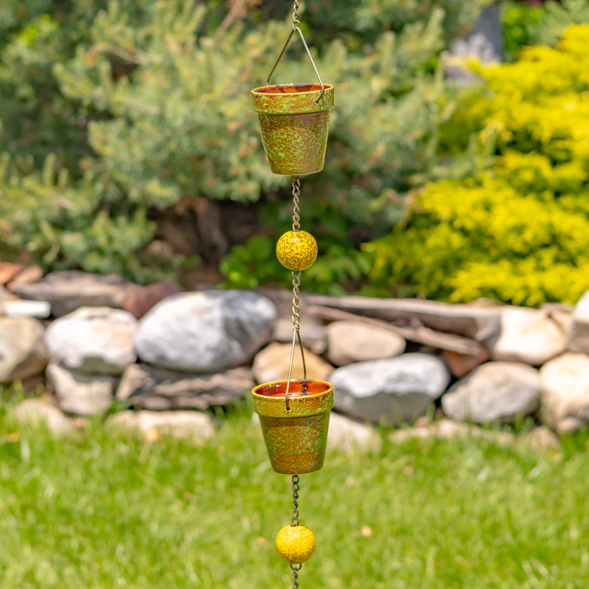 Two green bucket cups and yellow beads hang on a rain chain in a garden setting.
