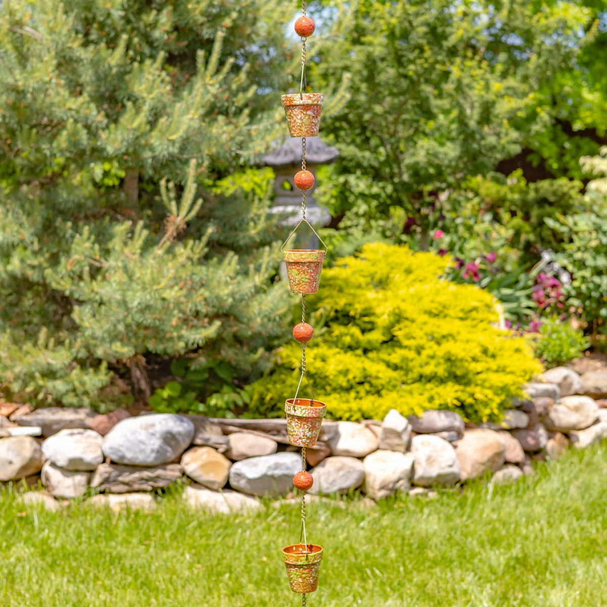 Rain chain with patterned cups and red beads hanging in a garden above a stone border.