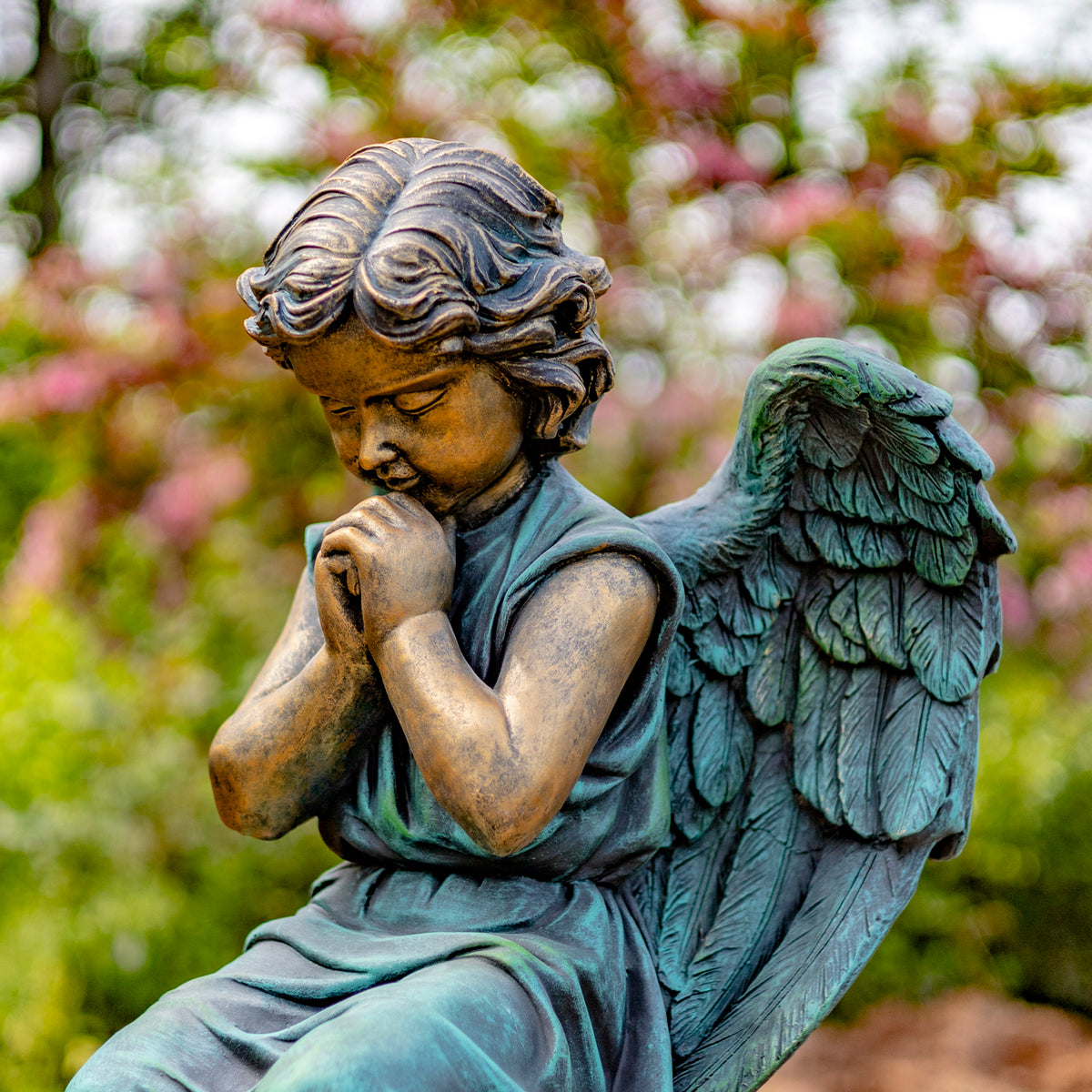 Seated angel statue with folded hands and a feathered wing, shown outdoors against blurred flowering greenery.