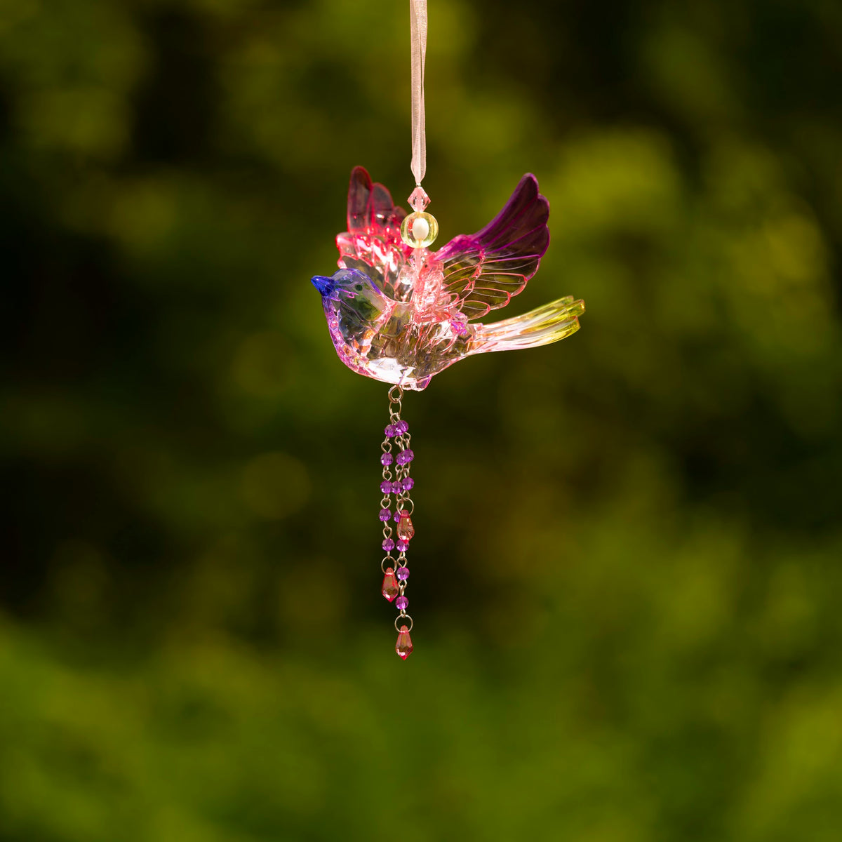 Colorful translucent bird ornament hanging against a softly blurred green background, with a beaded tail.