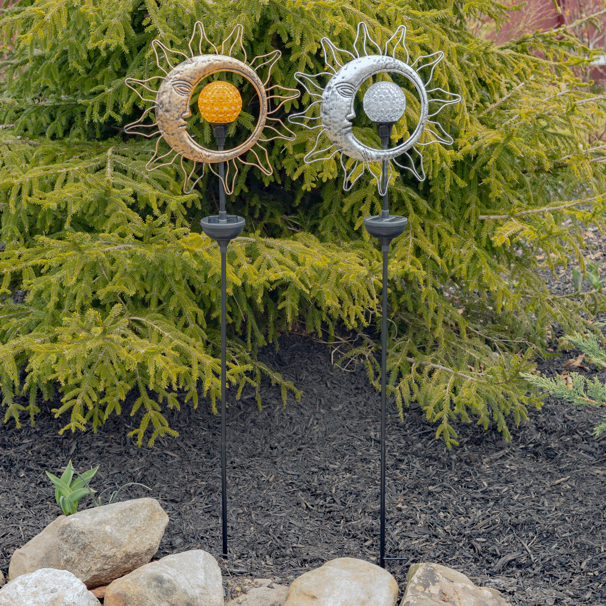 Two metal sun-and-moon garden stakes, one gold and one silver, standing among evergreen shrubs and rocks.