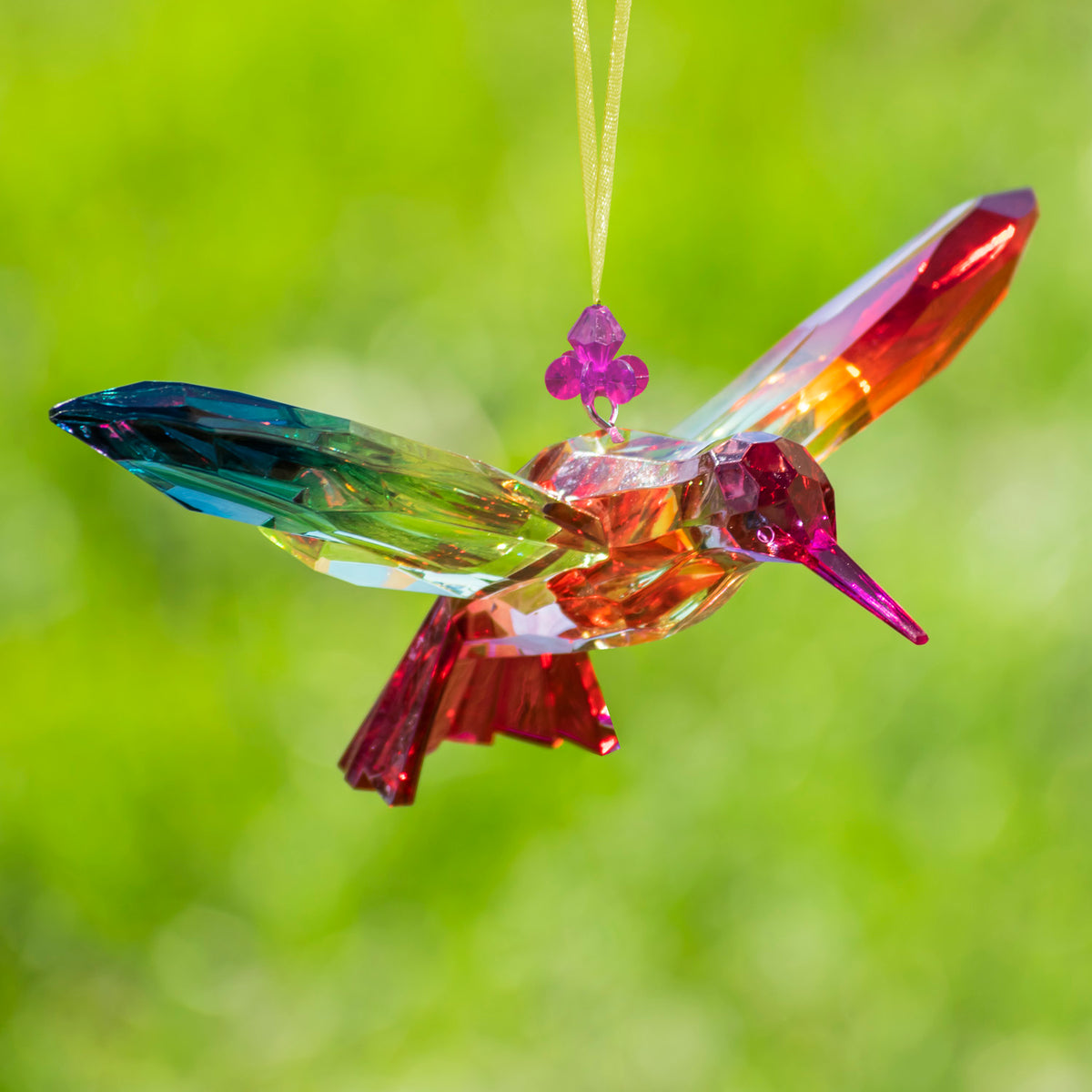 A multicolored faceted hummingbird ornament hangs by a pale cord, with wings spread and its beak pointing right.