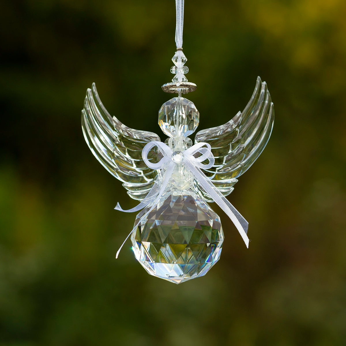 Clear faceted angel ornament with raised wings and a ribbon bow, hanging against a softly blurred green background.