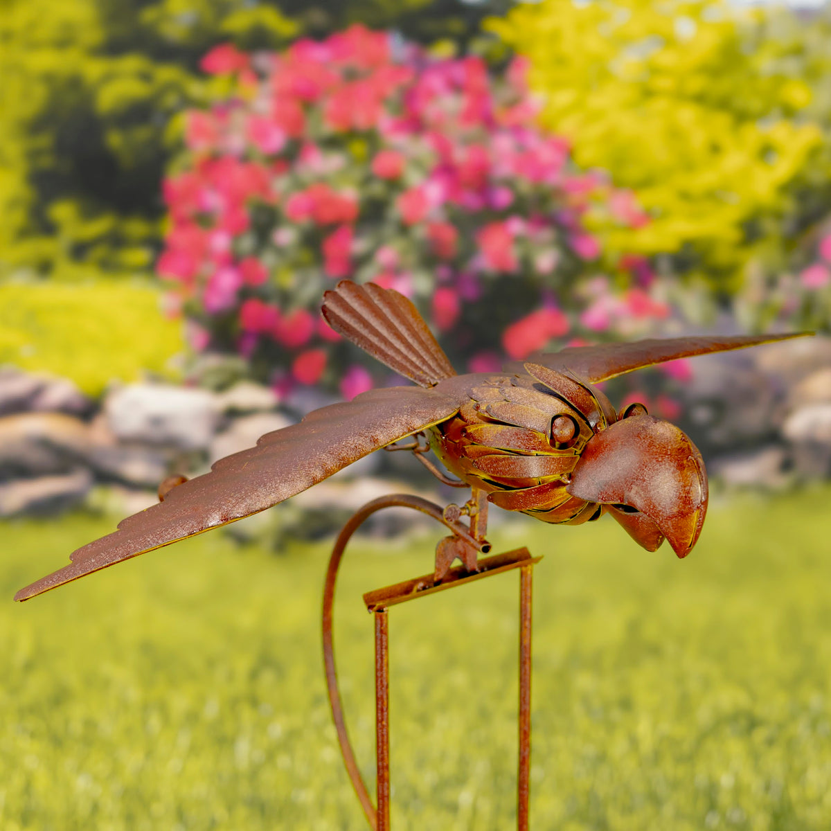 Rust-colored metal bird stake with outstretched wings in a garden with pink flowers and greenery.