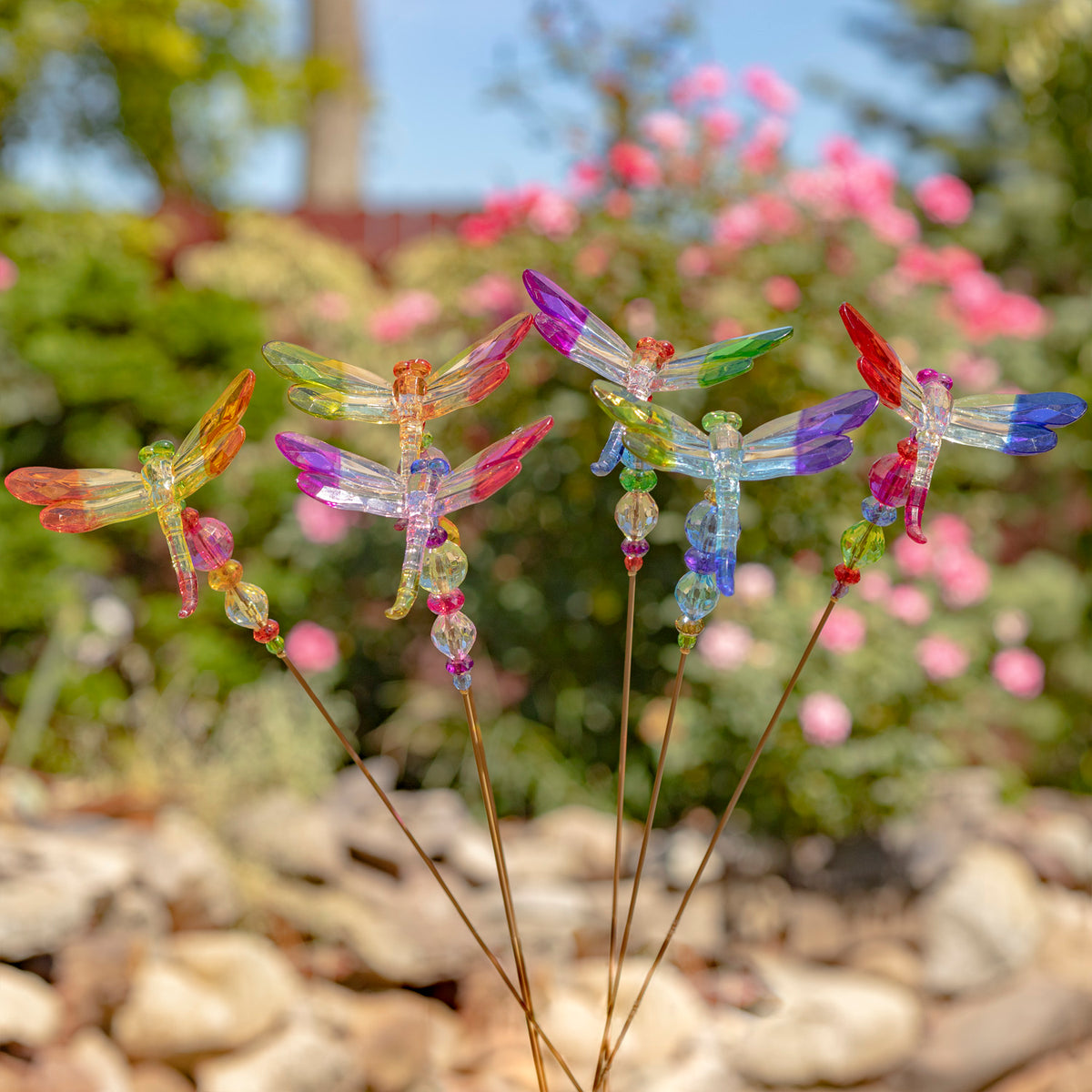 Several colorful dragonfly stakes stand together among garden flowers and rocks.