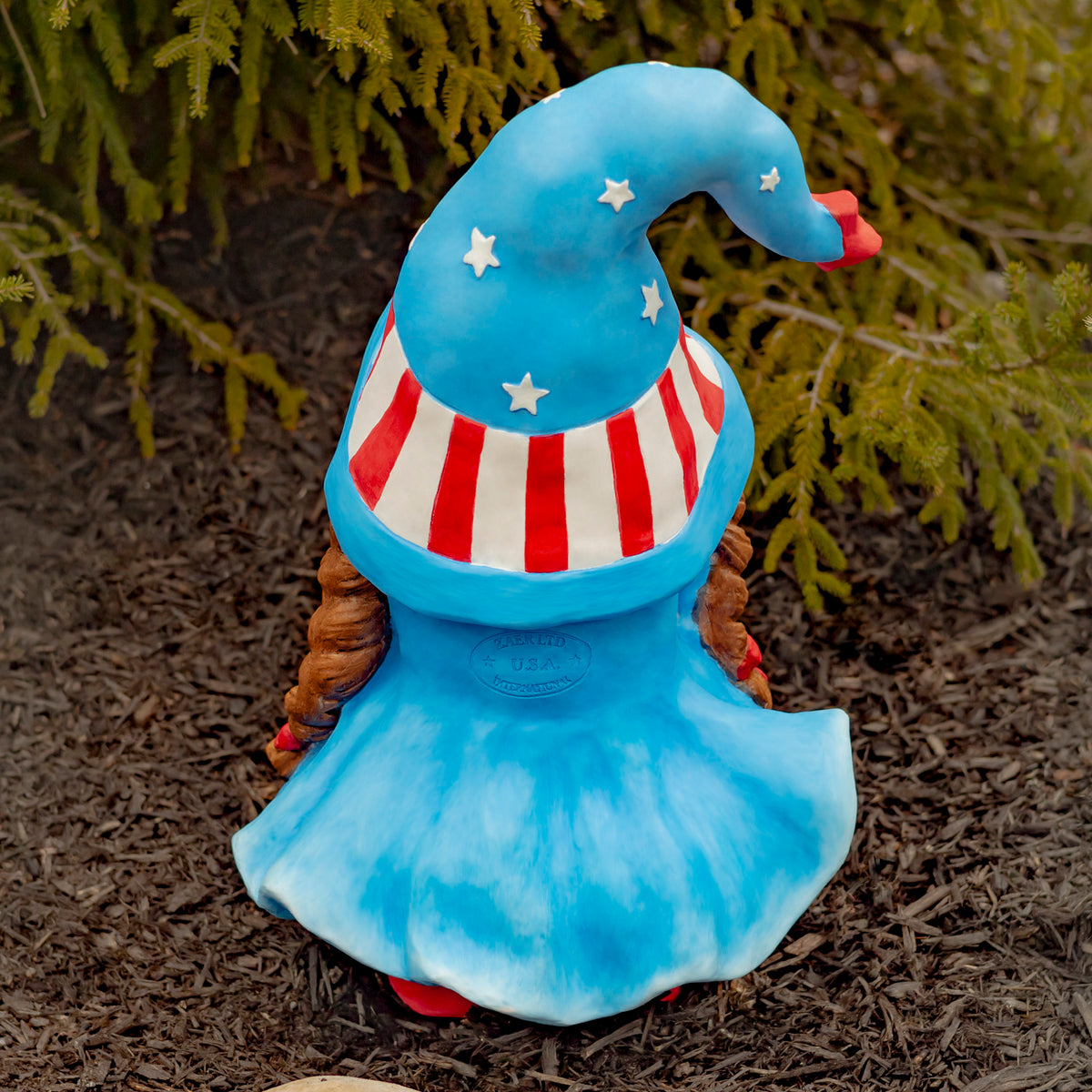 Back of the garden gnome, showing its curved starry hat, striped band, braids and blue dress.