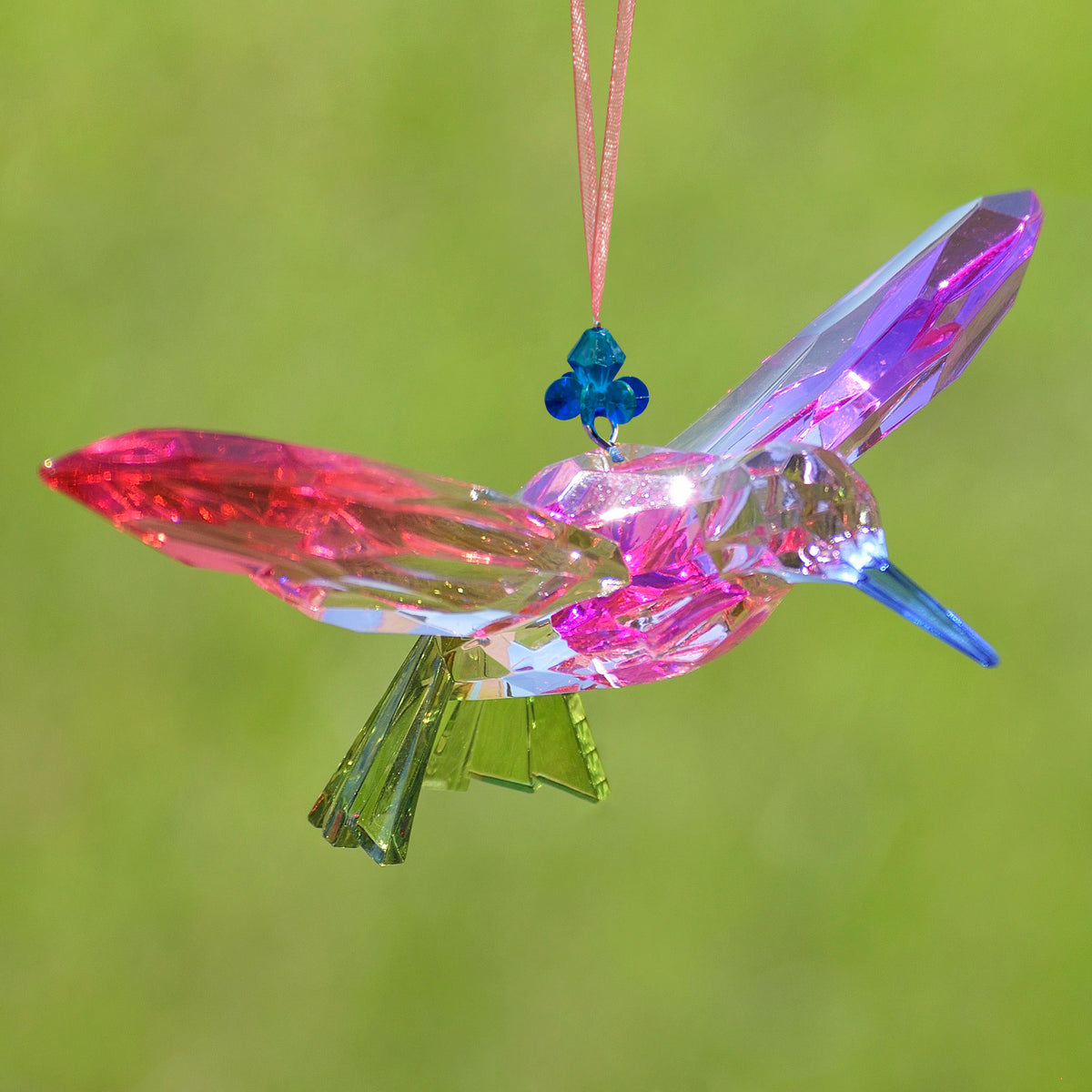 Pink and purple faceted hummingbird ornament suspended against a soft green background, with a blue beaded connector.