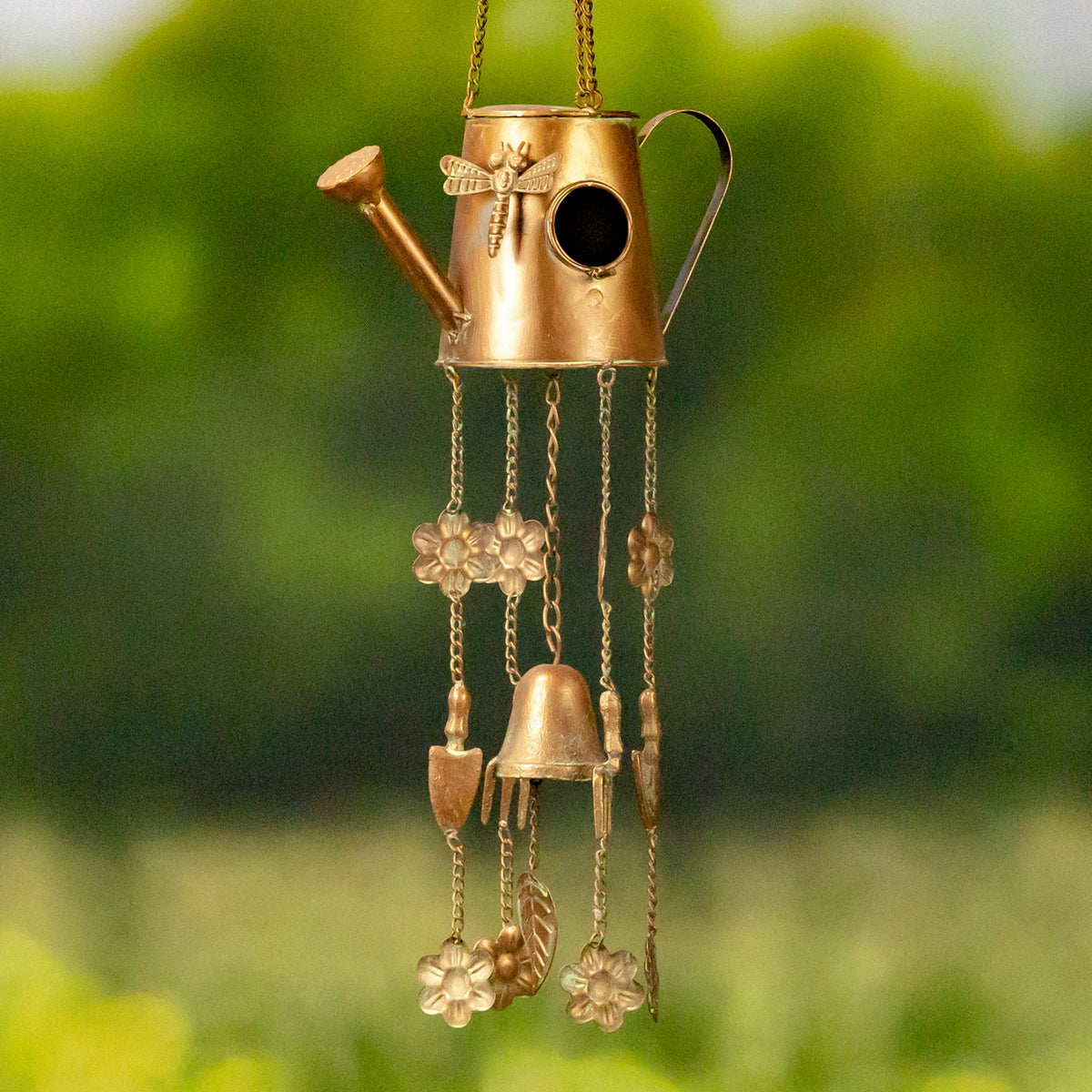 Antique-gold watering-can birdhouse wind chime with dangling flower and bell ornaments against a green background.
