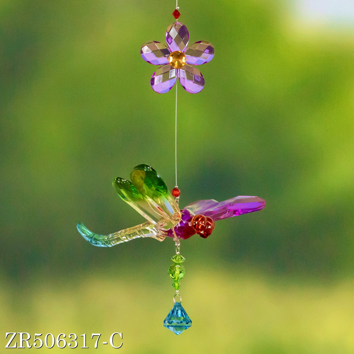 A jewel-toned dragonfly ornament hangs below a purple flower against a blurred green background.
