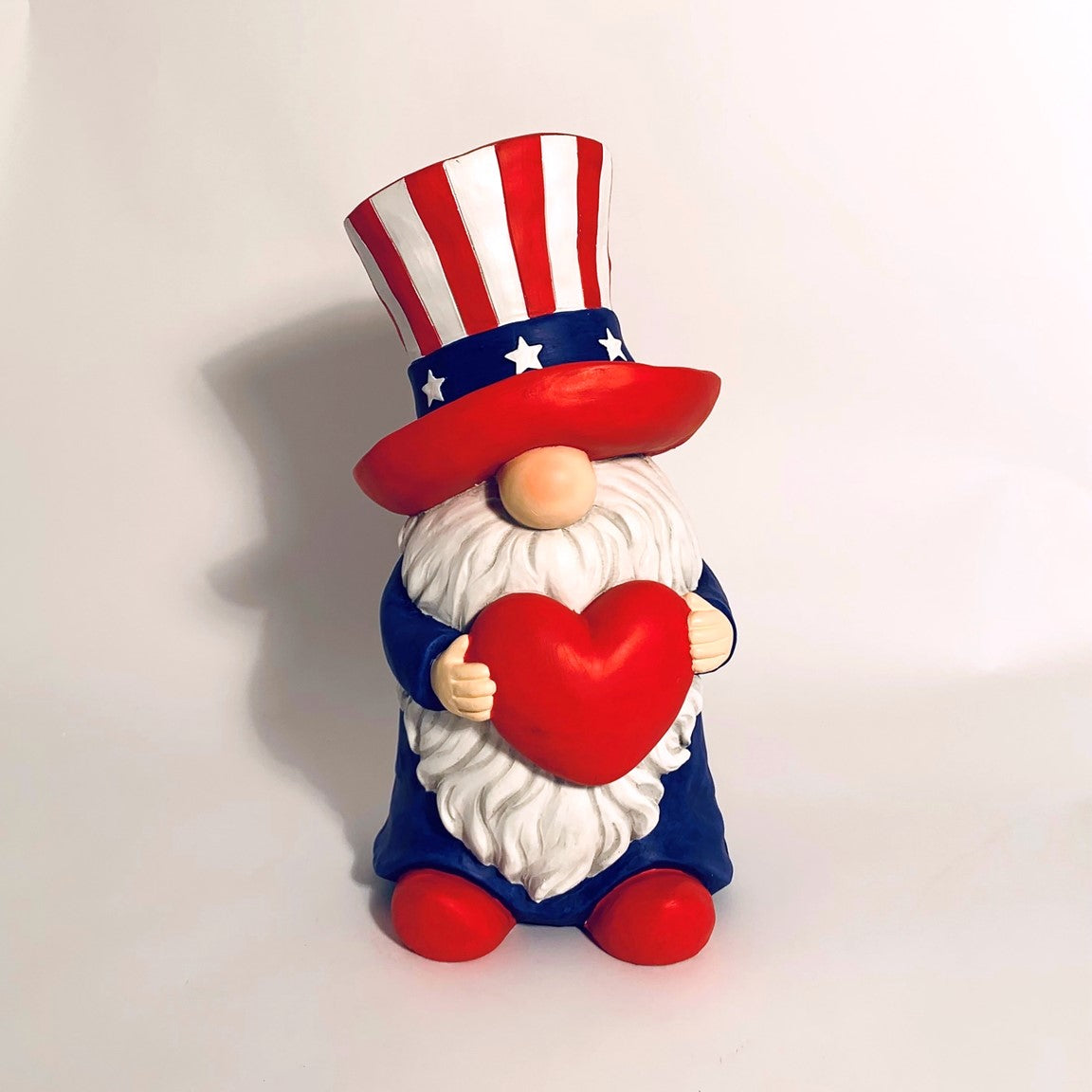 Front view of a red, white and blue gnome holding a red heart against a light backdrop