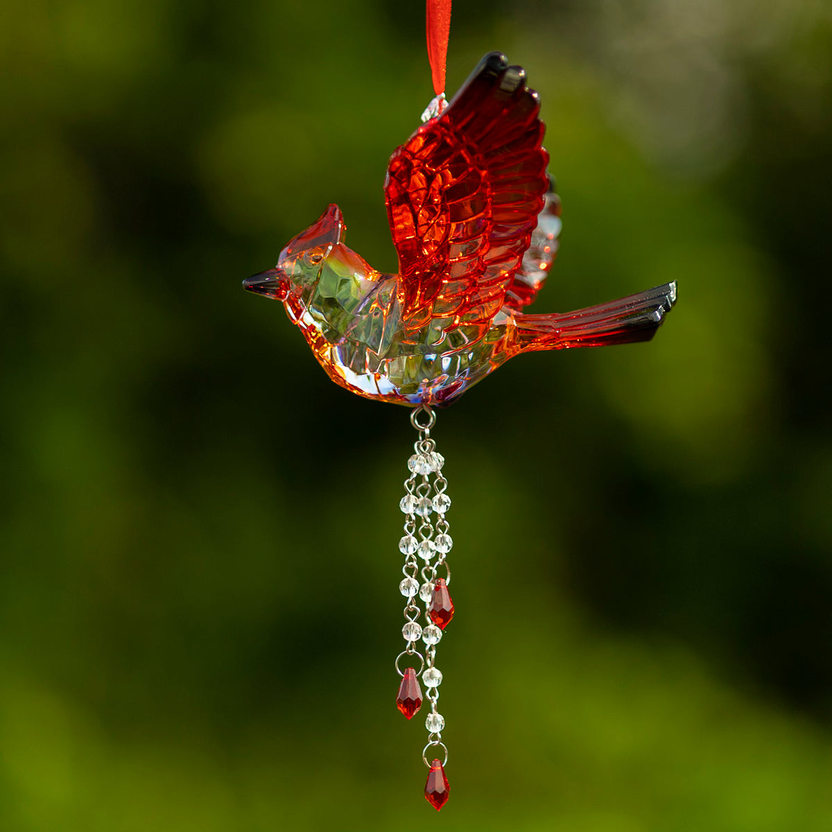 Faceted clear and red bird ornament with a raised wing and dangling beaded tassel against a green background.