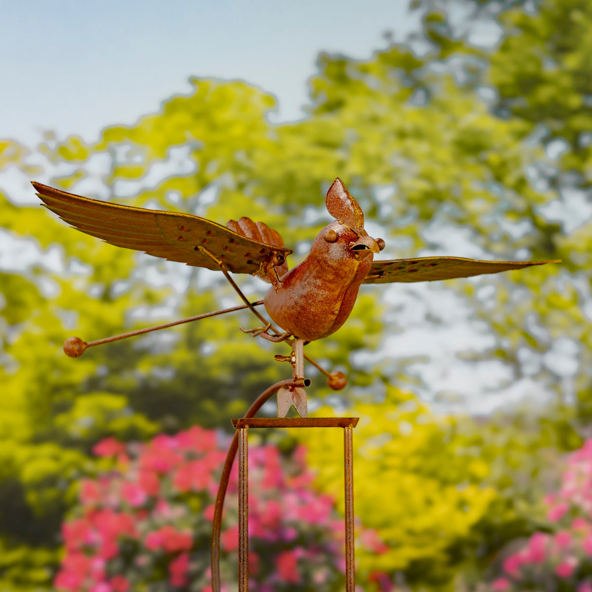Rust-colored metal bird with outstretched wings perched on a rocking stake in a flower garden.