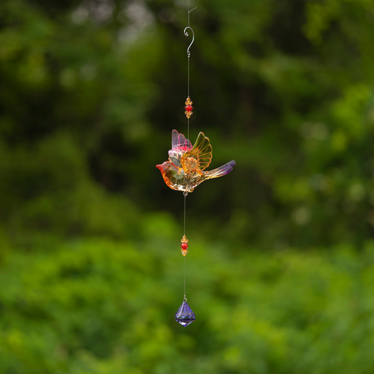 Acrylic bird ornament hangs from a long wire with red and gold beads and a purple faceted drop against greenery.