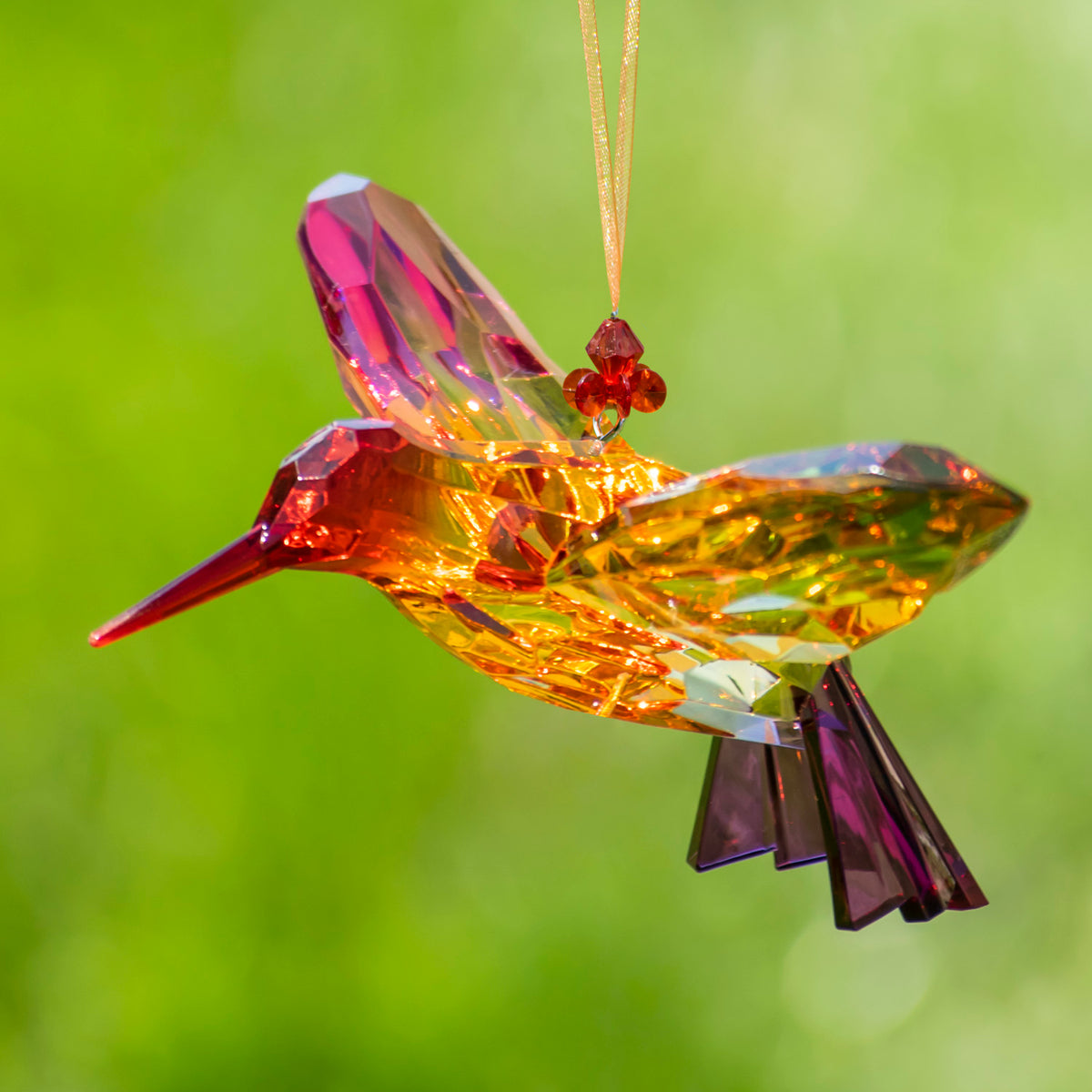 An orange and pink faceted hummingbird ornament hangs from a gold-colored cord, its beak pointing left.
