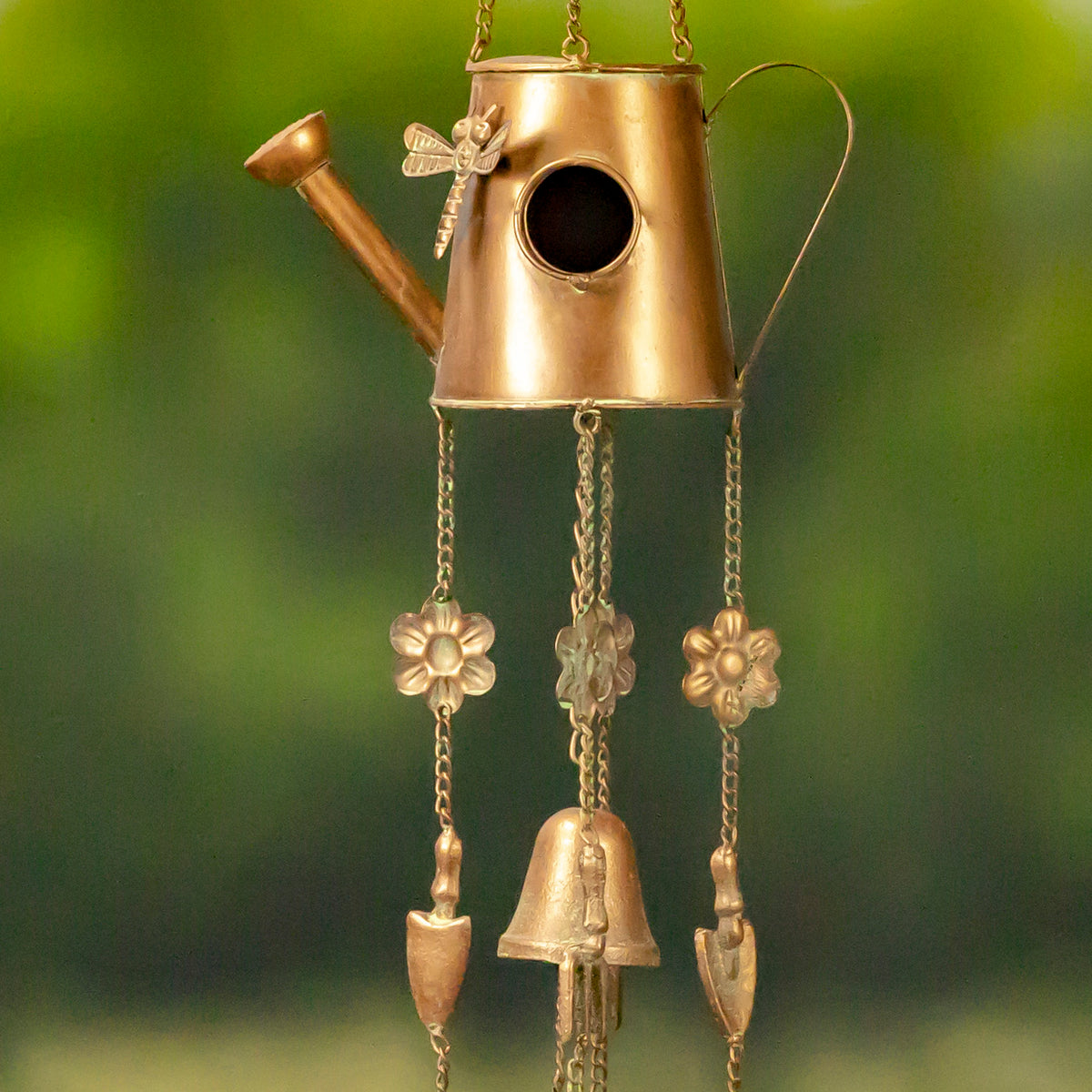 Closer view of the watering-can birdhouse, showing its round entrance, dragonfly accent, and hanging flower and bell
