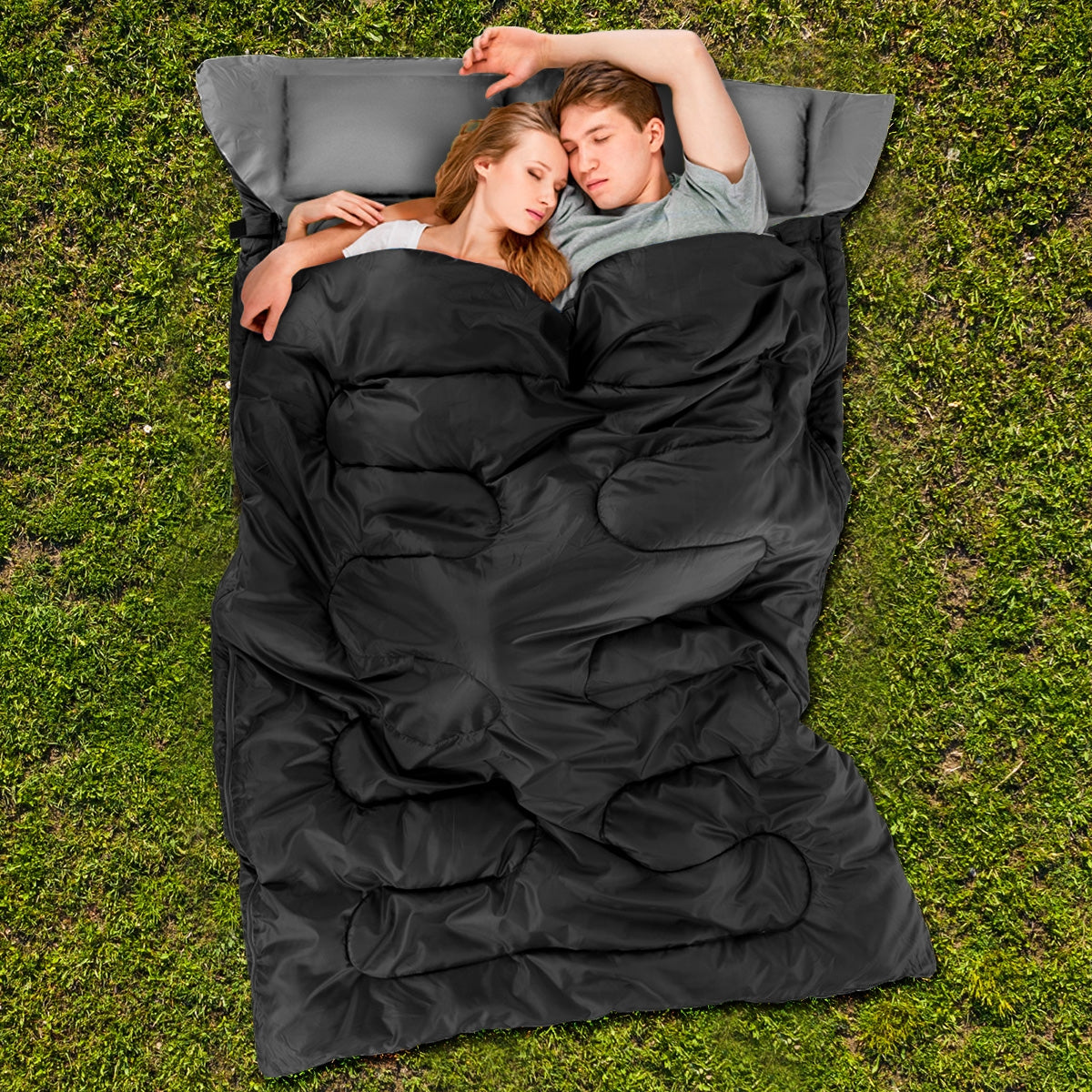 A couple lies together in an open black sleeping bag on grass, with gray pillows behind their heads.