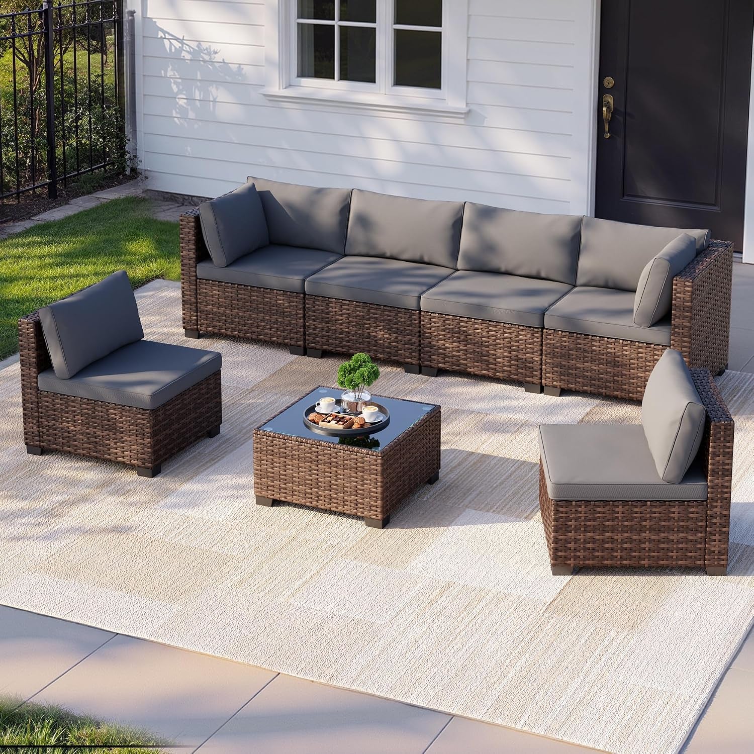 Outdoor patio scene showing a brown rattan sectional with grey cushions and a glass-top coffee table on a light rug.