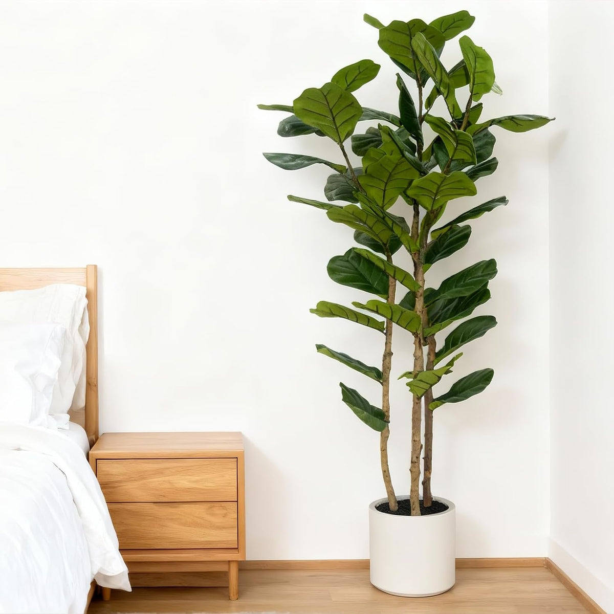 Tall artificial fiddle leaf fig in a white pot placed beside a wood nightstand and bed in a minimalist bedroom corner.