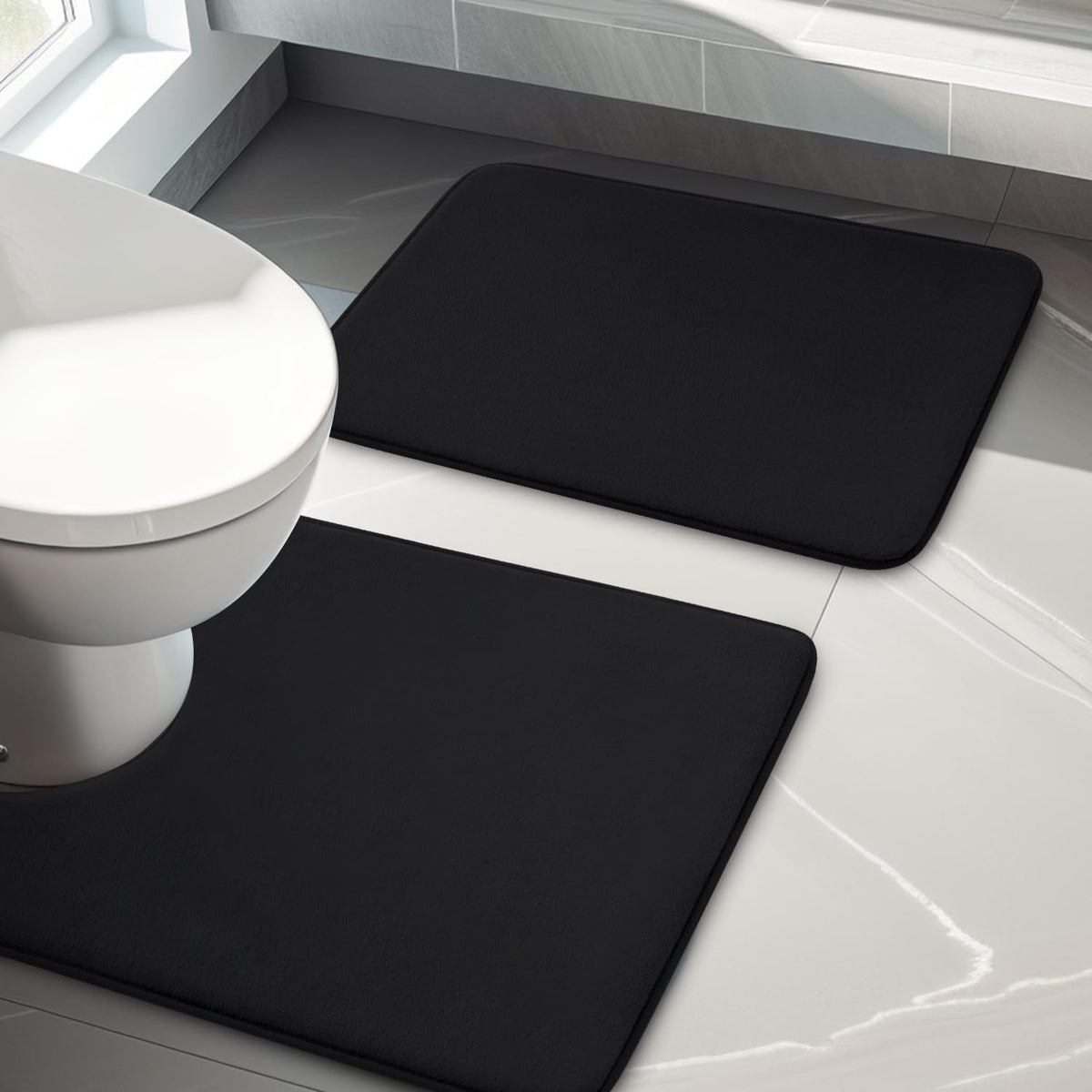 Overhead view of two black rectangular bath rugs beside a white toilet on glossy tile.