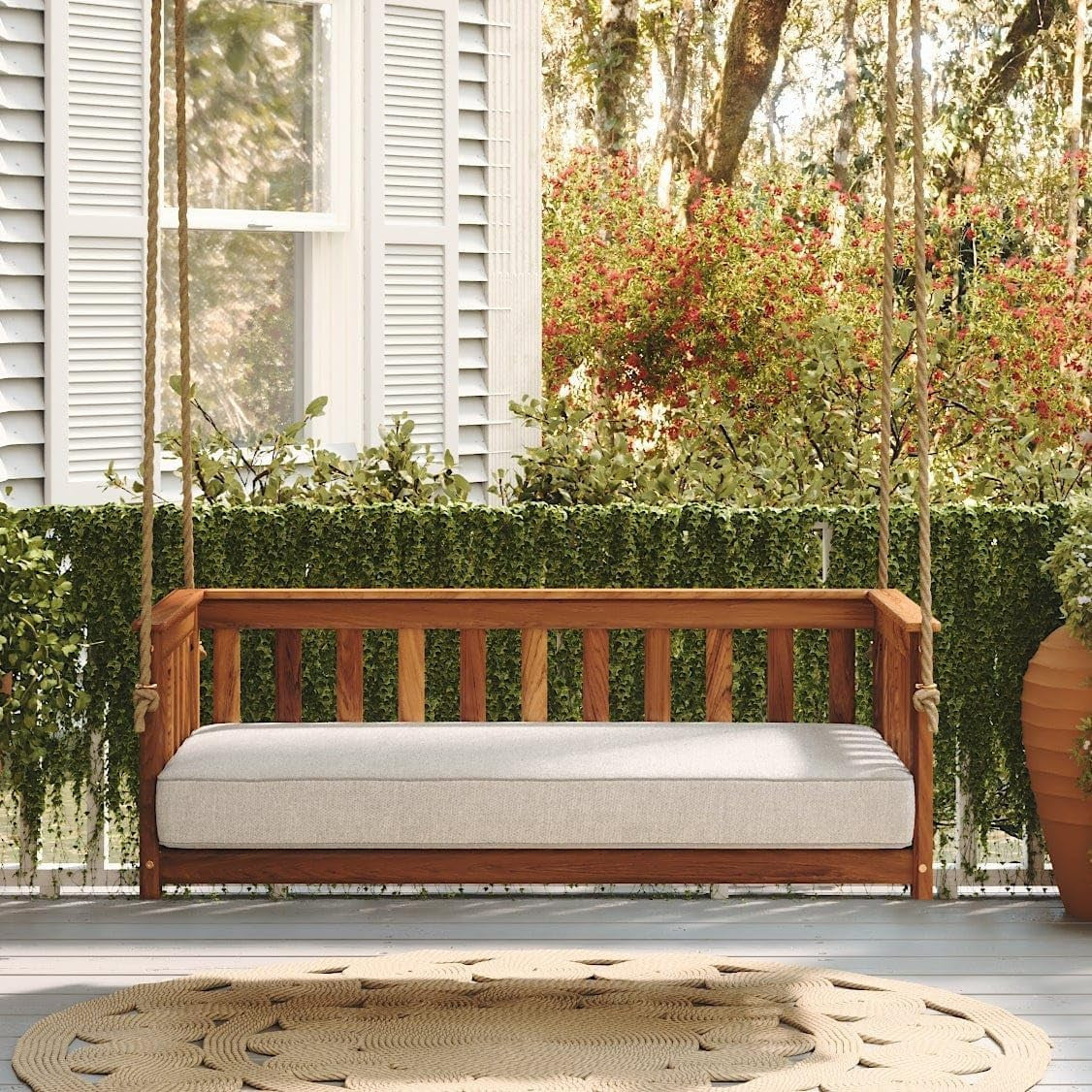 Front view of acacia porch swing with mattress cushion hanging on a porch with plants and greenery