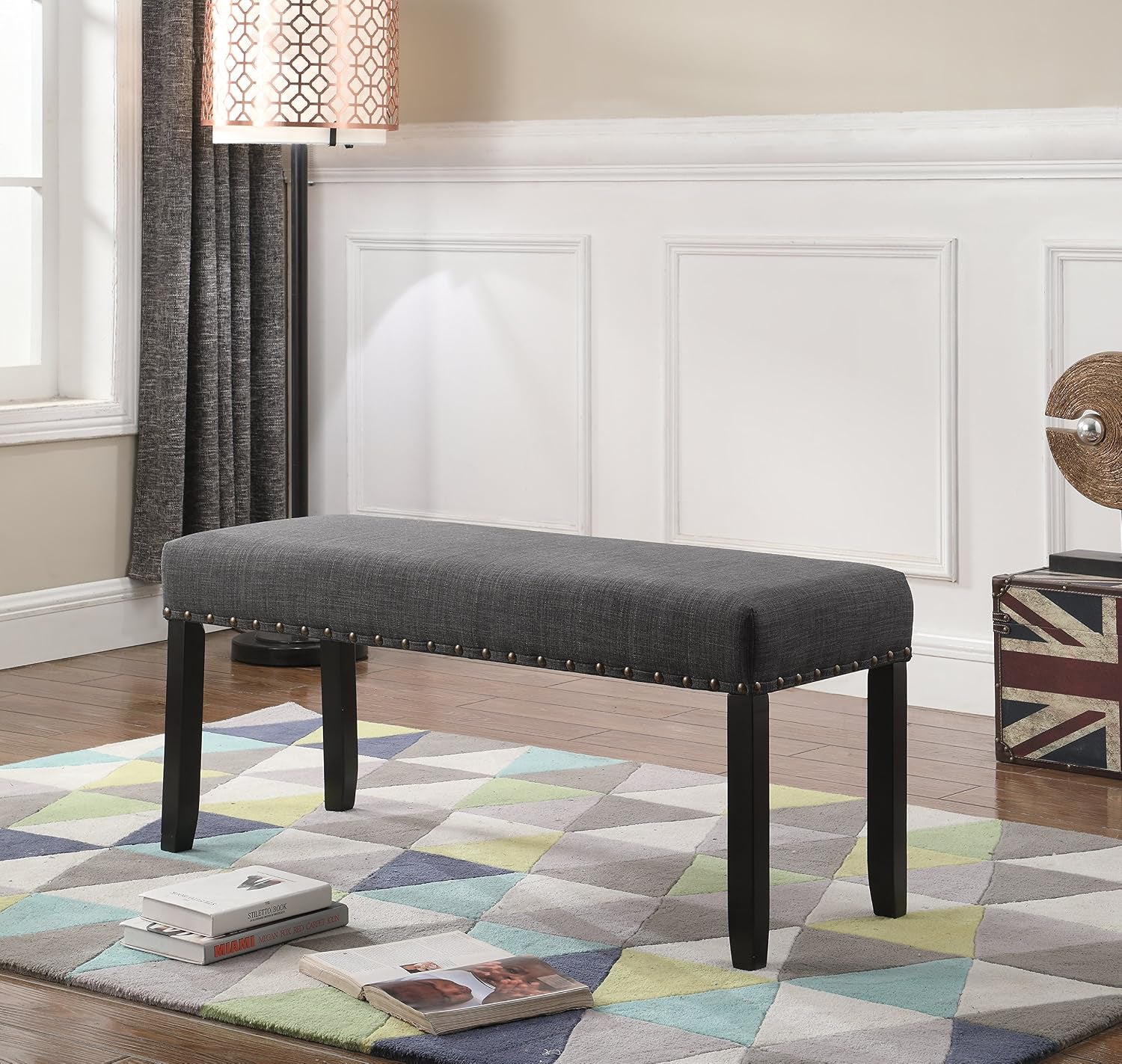 Full view of dark gray upholstered bench with nailhead trim placed on a geometric rug by a paneled wall